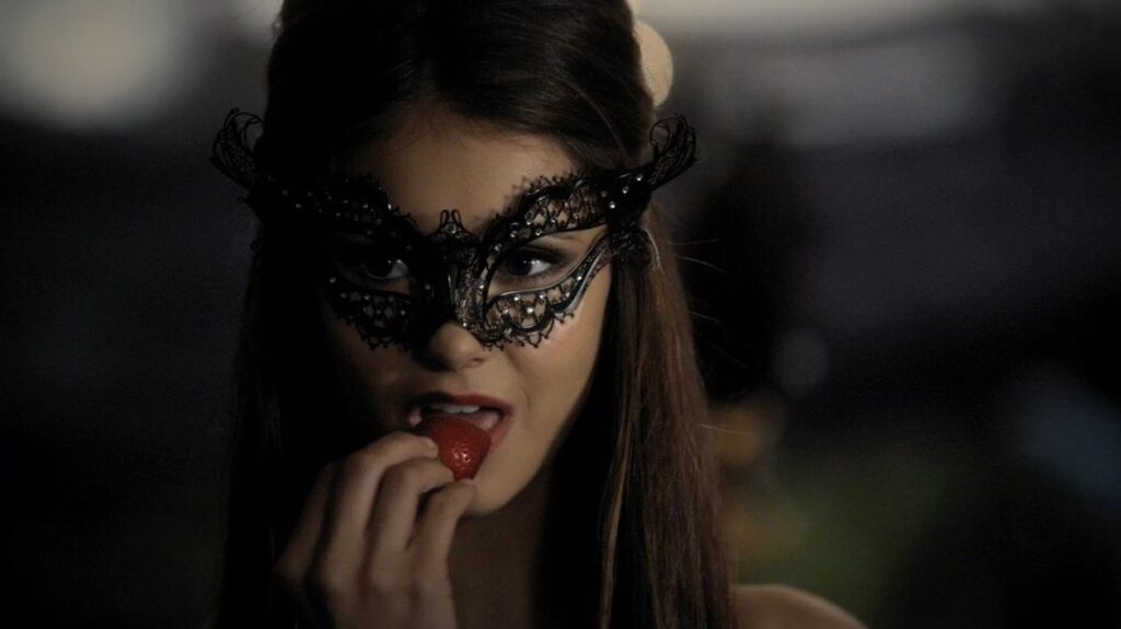 Nina Dobrev as Katherine Pierce with dark hair wearing an ornate black lace masquerade mask, holding a red strawberry to her lips with a seductive expression in a still from The Vampire Diaries.