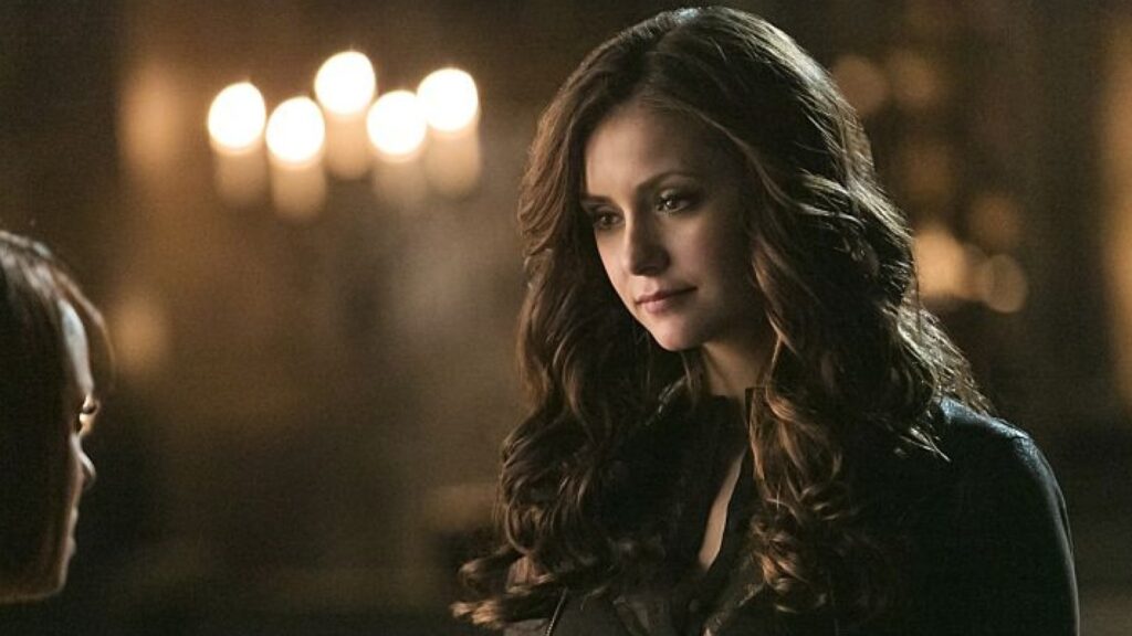 Nina Dobrev as Katherine Pierce with long dark wavy hair wearing a dark leather jacket, photographed in a dimly lit indoor setting with warm lighting in the background.