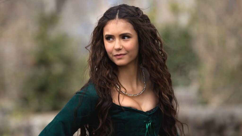 Nina Dobrev as Katherine Pierce with long curly dark hair wearing a dark green medieval-style dress with lace-up details and a silver necklace, standing outdoors.