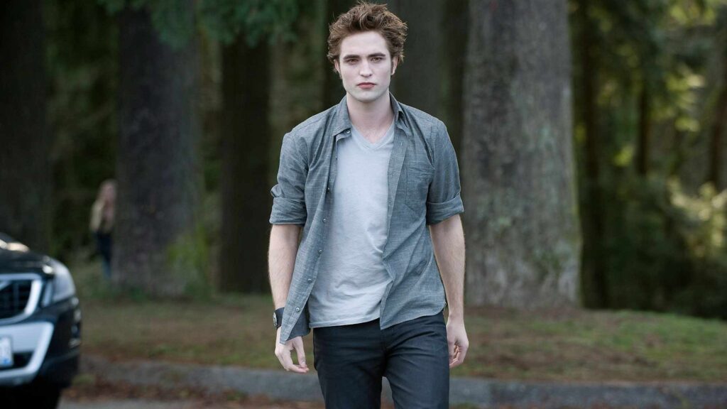 Edward Cullen (Robert Pattinson) walks confidently outdoors with trees in the background, wearing a gray shirt and showing his characteristic intense expression.