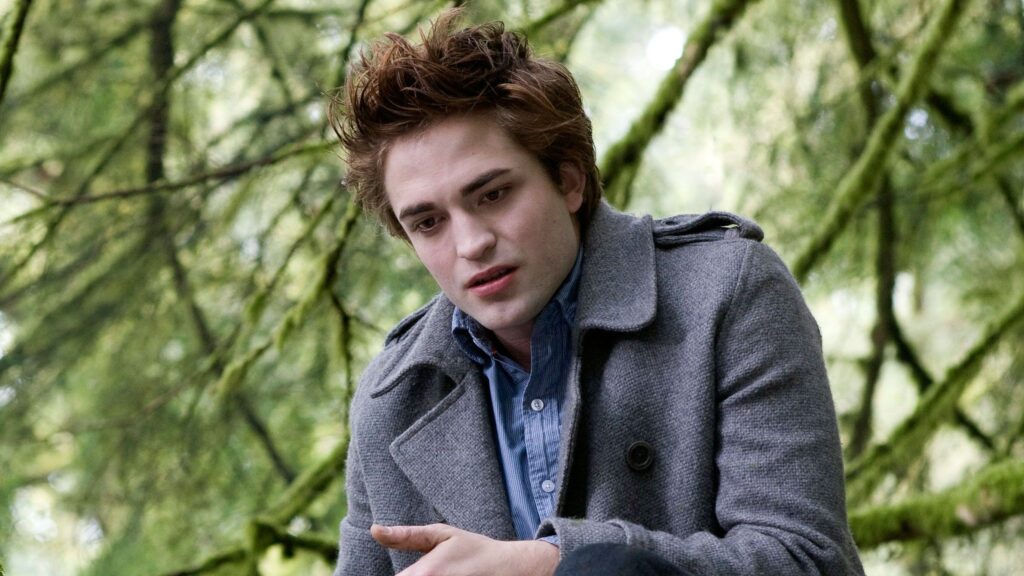 Edward (Robert Pattinson) wearing a gray coat in a forest setting, arms together, having a serious expression while talking to Bella (Kristen Stewart) off-screen during the reveal scene.