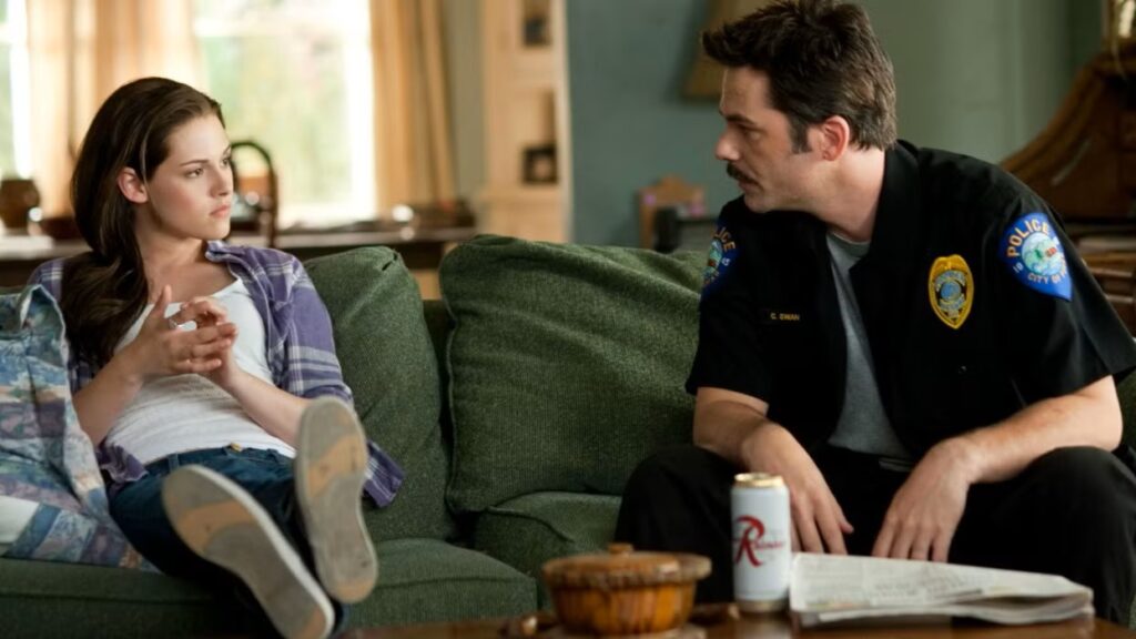 Bella Swan (Kristen Stewart) sits on a couch with her father Charlie (Billy Burke) who is wearing his police uniform, sharing a quiet moment together in their home.