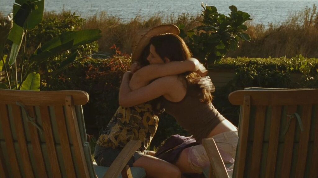Bella (Kristen Stewart) and her mother Renée (Sarah Clarke) embrace emotionally in an outdoor tropical setting with wooden chairs.