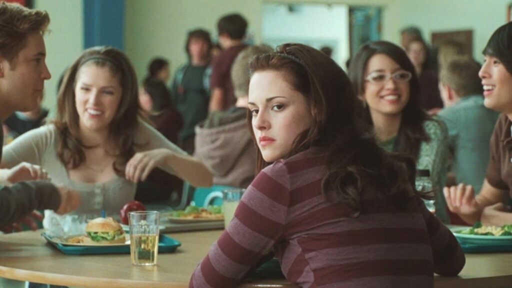 Bella Swan (Kristen Stewart) sits at the high school cafeteria table with her friends Mike Newton (Michael Welch), Jessica Stanley (Anna Kendrick), Angela Weber (Christian Serratos), and Eric Yorkie (Justin Chon), along with other students in the background.