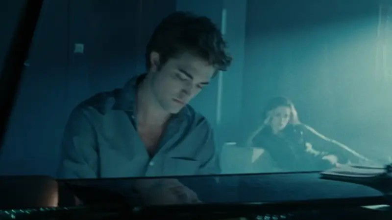 Edward Cullen (Robert Pattinson) sits at a piano in a dimly lit room with Bella (Kristen Stewart) visible in the background in a still from the first Twilight movie.