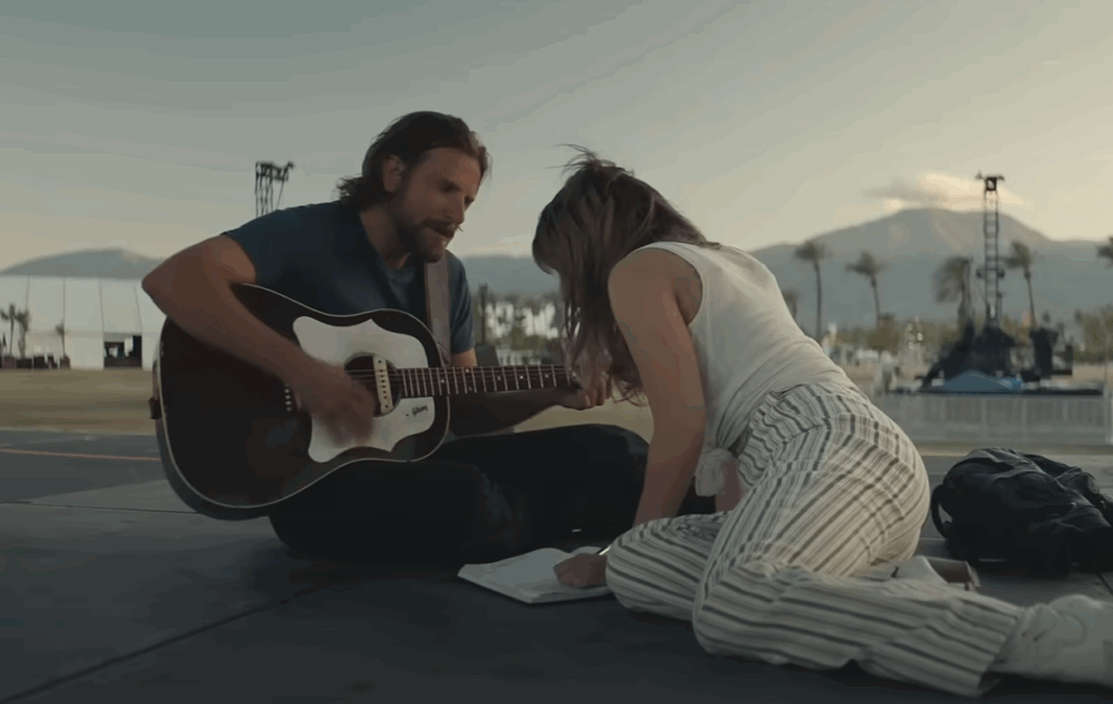 Bradley Cooper holding a guitar and sitting in front of Lady Gaga. 