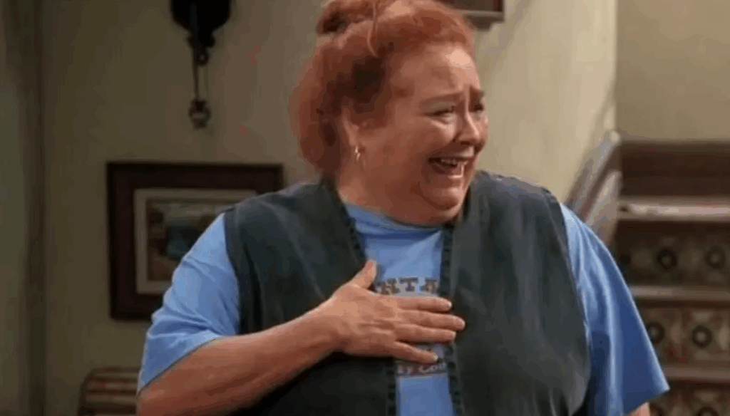 Berta is wearing a blue t-shirt and a vest. She is laughing heartily while holding her hand to her chest in the show Two and a Half Men.