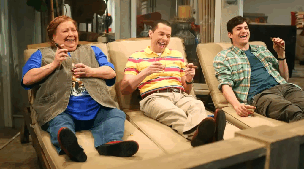 Berta is sitting on a lounge chair with Alan and Walden. They are all laughing and enjoying cigars and drinks on a patio or in a living area.