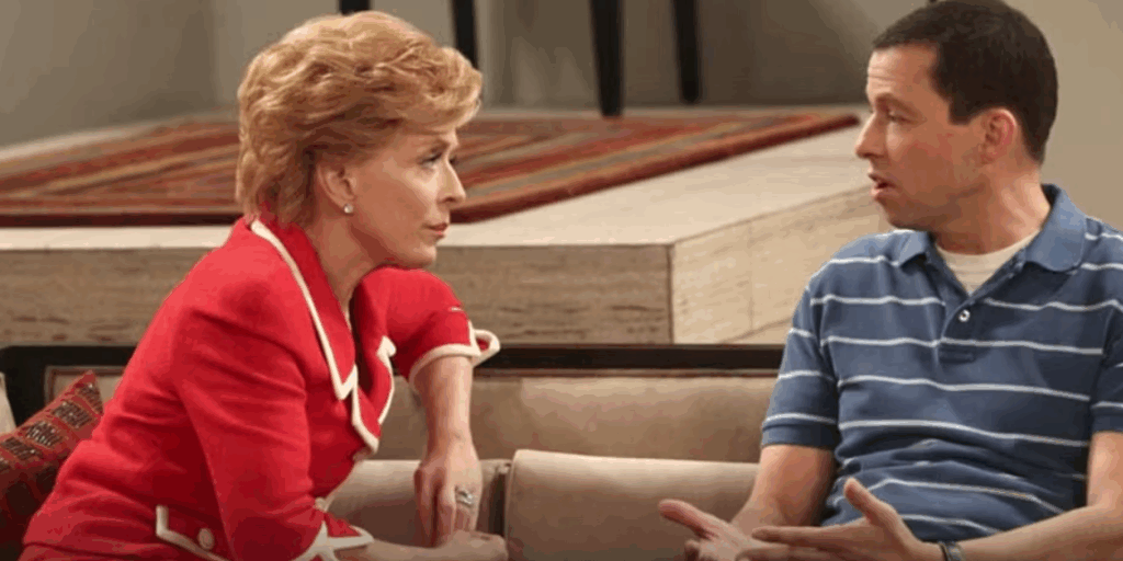 Evelyn Harper is sitting on a couch in a living room, wearing a bright red jacket. She is having a discussion with Alan.