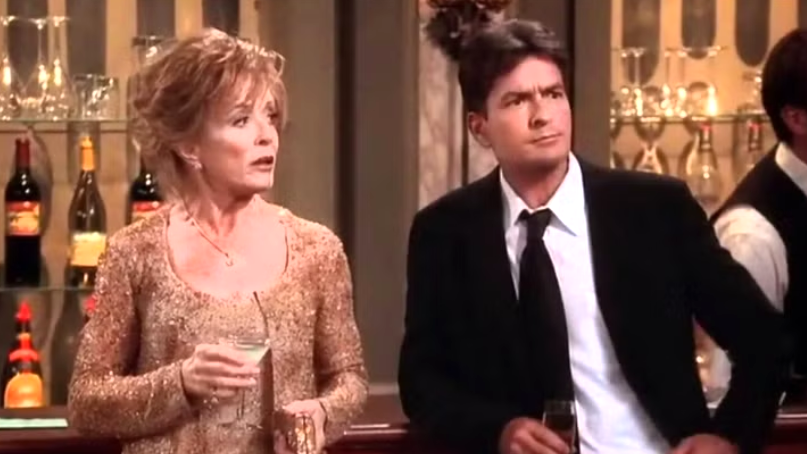 Evelyn Harper with short, blonde hair, wearing a shimmering golden dress, stands at a bar holding a drink. She is talking to Charlie.