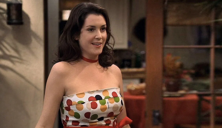 Rose with brown hair is wearing a low-cut dress with a colorful polka-dot pattern. She is smiling. She has a red ribbon tied around her neck and another tied at her waist.
