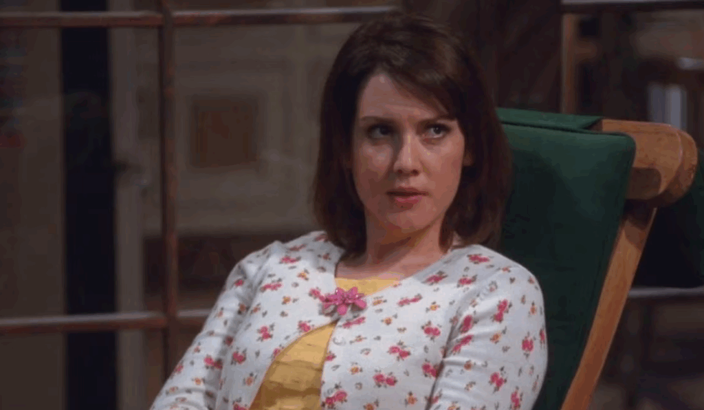 Rose is sitting on a chair, wearing a white cardigan with a pink floral print and a yellow shirt underneath.