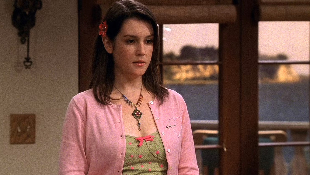 Rose is wearing a light pink cardigan over a green top, with a red flower in her hair and a necklace with a large pendant. She looks serious and is standing in front of a glass door.