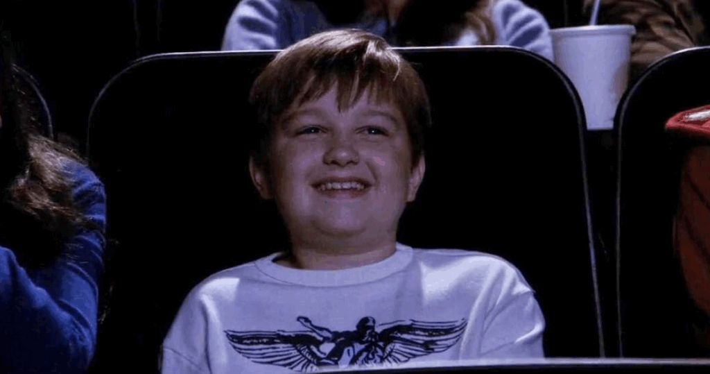 Jake is at a movie theater, sitting in a dark chair and laughing with a wide smile on his face.