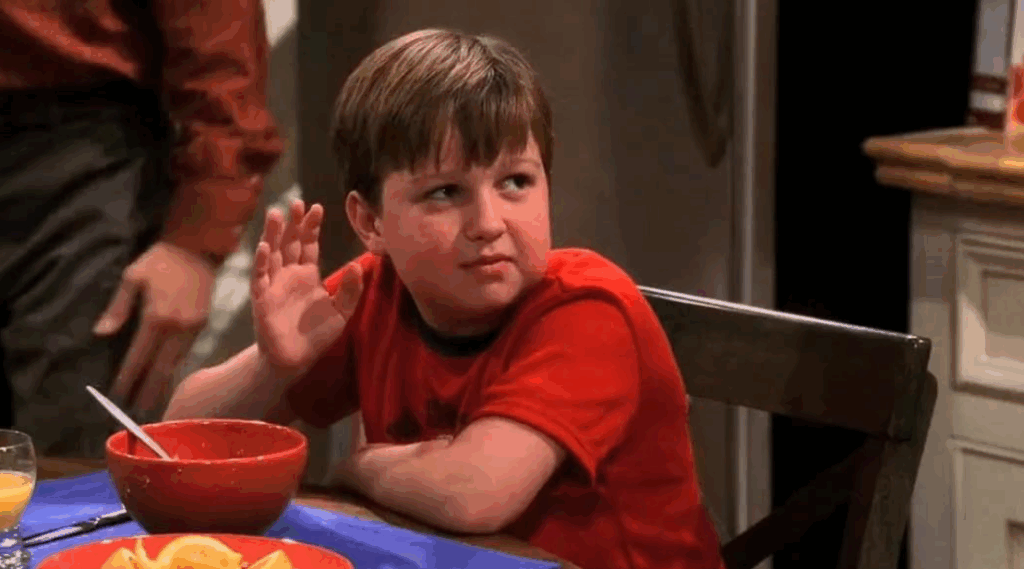 Jake in a red t-shirt, is sitting at a table with a bowl of cereal. He is holding up one hand.