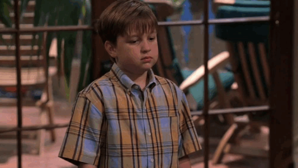 Jake is standing, looking down with a sad expression, wearing a blue and yellow plaid short-sleeved shirt.