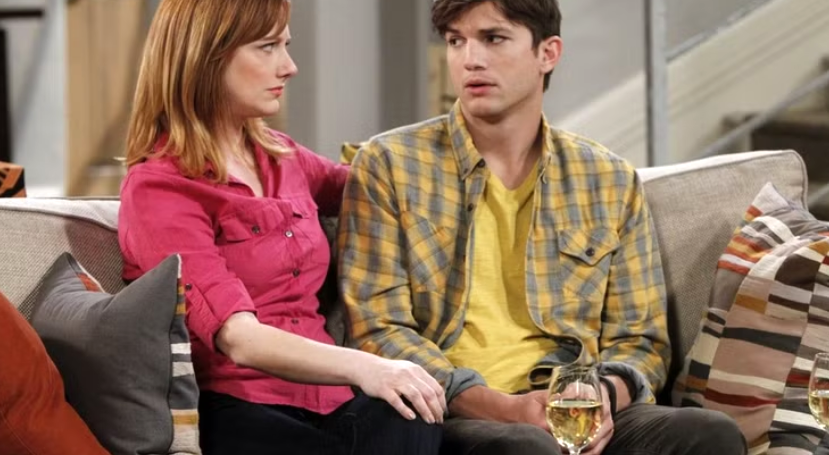 Walden Schmidt is sitting on a couch with a woman, holding a glass of wine and looking to the side.