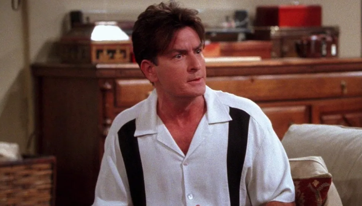 Charlie is sitting on a couch, wearing a white bowling shirt with black stripes on the front and looking to the side.
