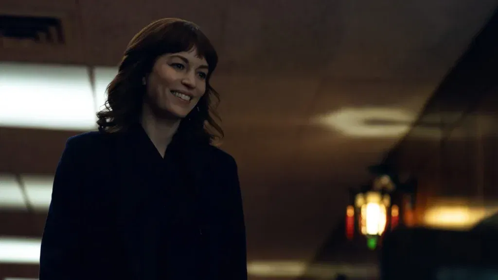 Britt Lower as Helly R in a still from Severance wearing a dark outfit and smiling while walking through dimmed office corridors.