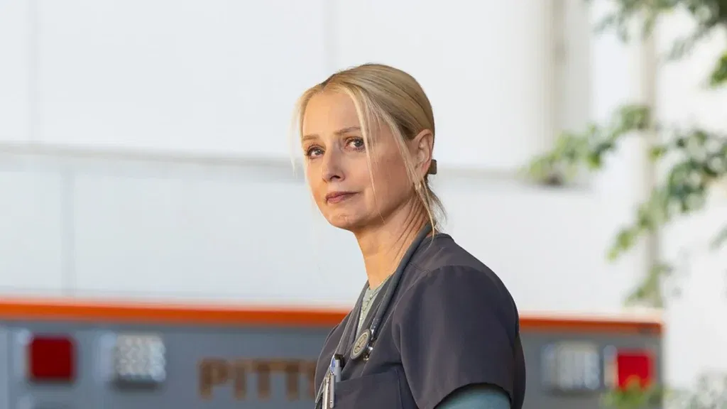Katherine LaNasa as Dana Evans in The Pitt wearing her medical scrubs with a stethoscope around her neck and hair tied back is looking over her shoulders towards the camera with a contemplative expression.