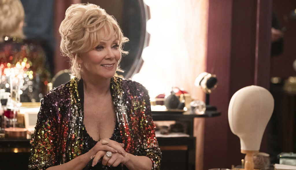 Jean Smart as Deborah Vance with blonde curly hair wearing a sequined or beaded jacket in multiple colors (appearing to have gold, red, and other metallic tones), sitting in an upscale dressing room in a still from Hacks.