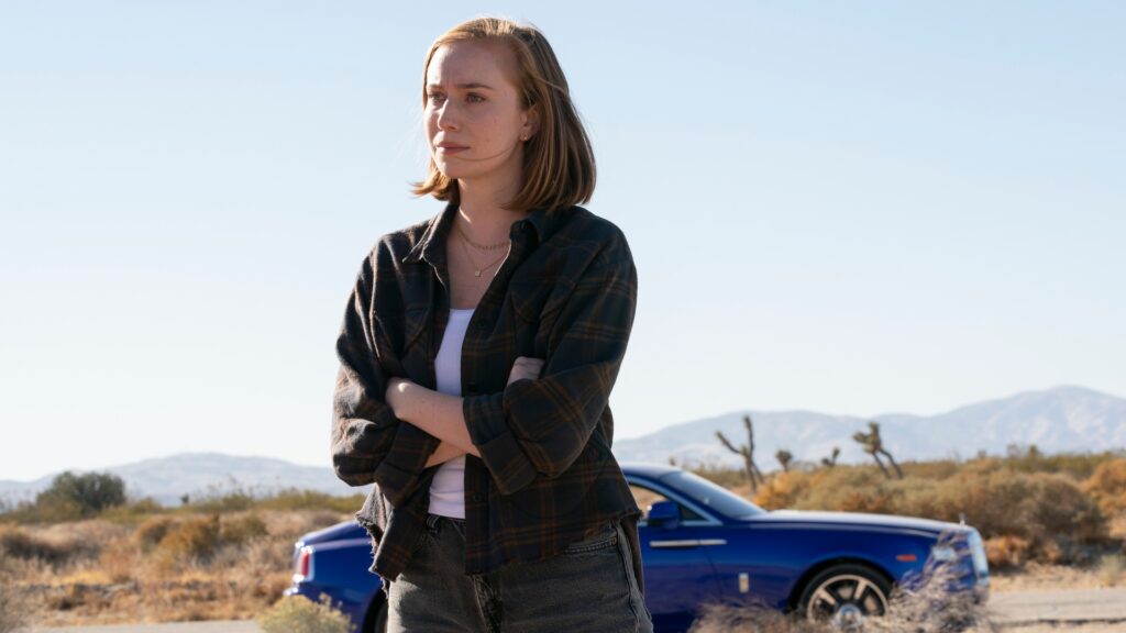 Hannah Einbinder as Ava Daniels with shoulder-length blonde hair wearing a dark jacket, standing outdoors in a desert landscape with mountains in the background and a blue car visible in a still from Hacks.