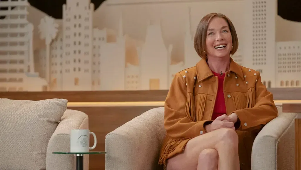 Julianne Nicholson as Dance Mom with shoulder-length brown hair wearing a tan/camel colored suede jacket with fringe details and studded embellishments, sitting on a light-colored couch in a still from Hacks.