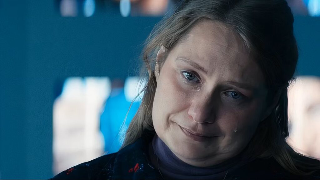 Merritt Wever as Gretchen George with blonde hair wearing a dark winter coat or jacket, photographed in dramatic lighting with a serious expression while tearing up in a still from Severance.