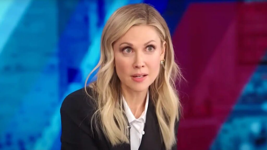Desi Lydic in professional attire (black blazer and white shirt) speaking on The Daily Show: Desi Lydic Foxsplains, with colorful blue and red lighting in the background.