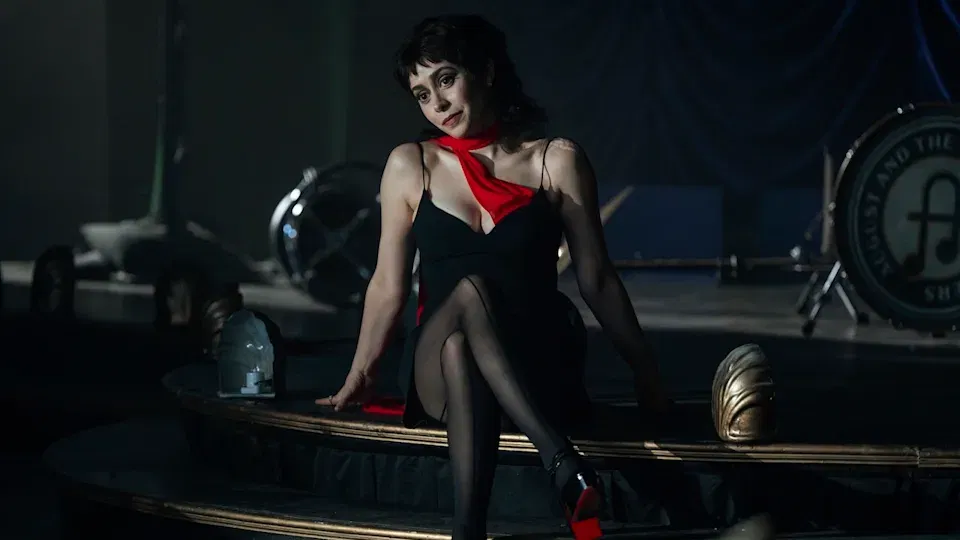 Cristin Milioti as Sofia Falcone in The Penguin with short dark hair wearing a black dress with a red scarf or collar detail, sitting on a wooden staircase in a dimly lit, vintage-style room.