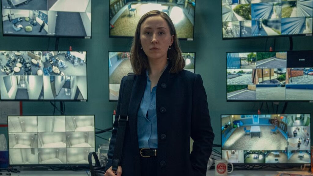 Erin Doherty as Briony Ariston in Adolescence with shoulder-length brown hair wearing a dark blazer and light blue shirt, standing in a monitoring room with multiple screens displaying surveillance footage.