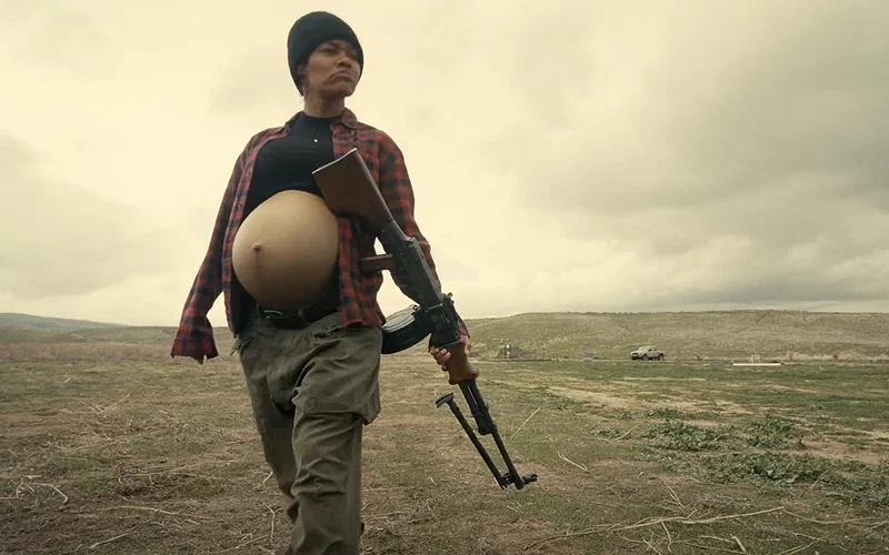 Teyana Taylor wearing a beanie and holding an AR in a field in One Battle After Another.