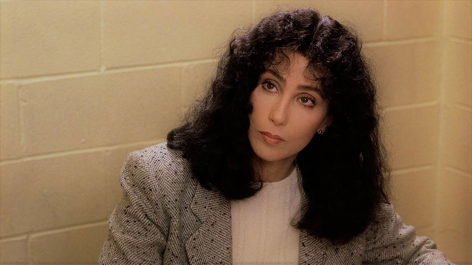 Cher in the 1980s with voluminous dark curly hair, wearing a light-colored sweater and sitting casually in a still from the movie Suspect.