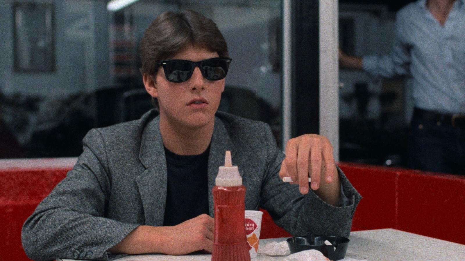 Tom Cruise in Risky Business sitting at a diner table wearing sunglasses and a gray blazer, with ketchup and condiments visible on the red booth table.