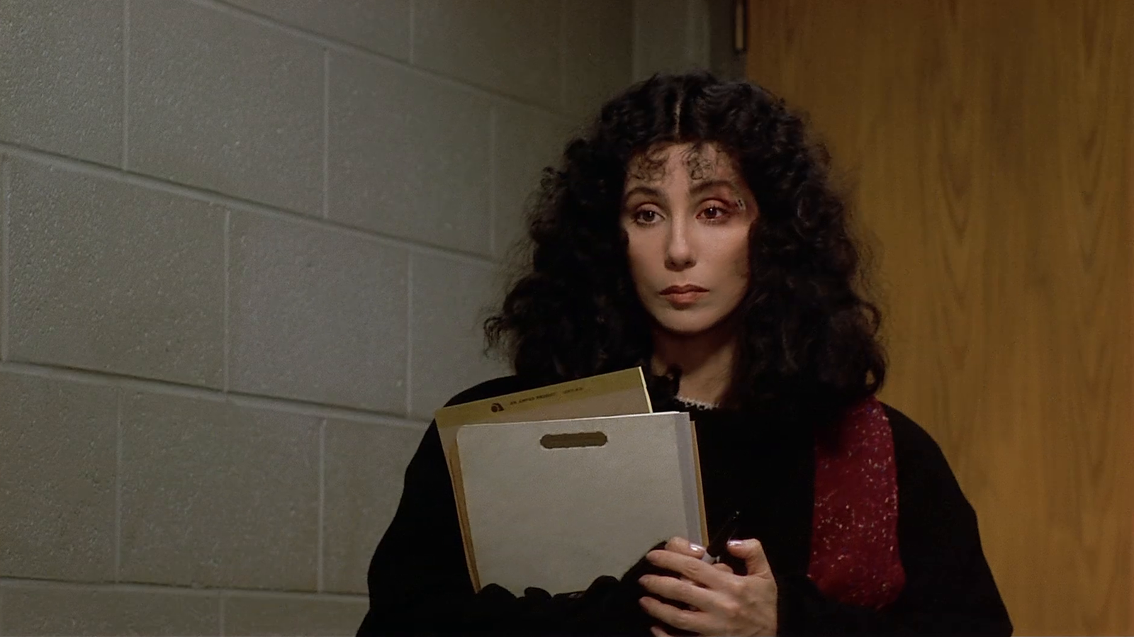 Cher in the 1983 film Suspect, wearing dark clothing and holding folders, looking serious while standing in an indoor setting.