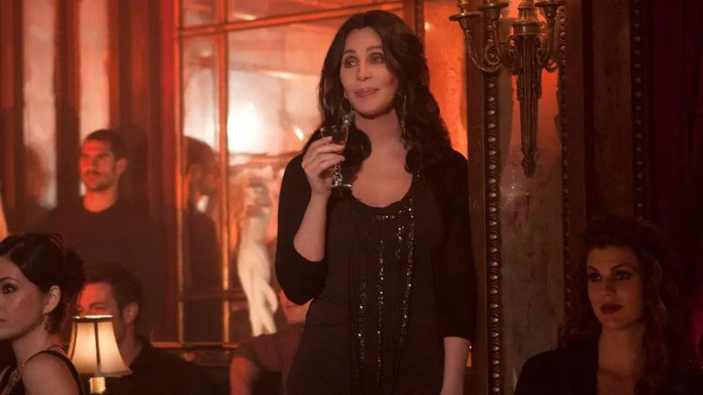 Cher in an elegant black sequined dress, holding a champagne flute in an intimate venue setting from the movie Burlesque.