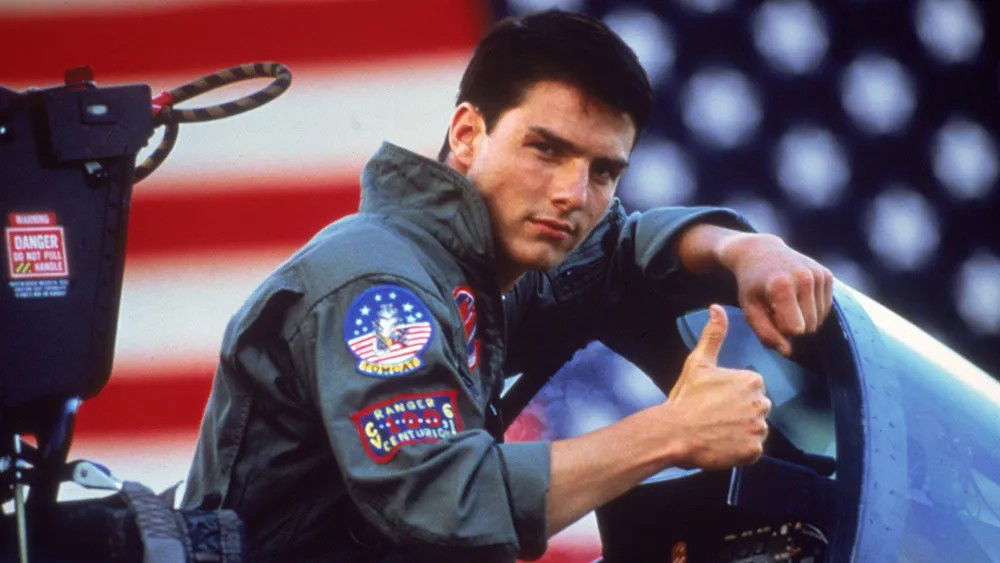 Tom Cruise as Maverick in Top Gun, wearing his flight suit and giving a thumbs up gesture while standing next to military aircraft with an American flag in the background.