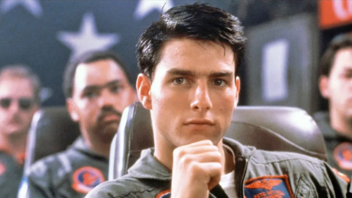 Young Tom Cruise in his iconic Top Gun role, wearing a military flight suit and sitting in an aircraft cockpit with a confident expression.