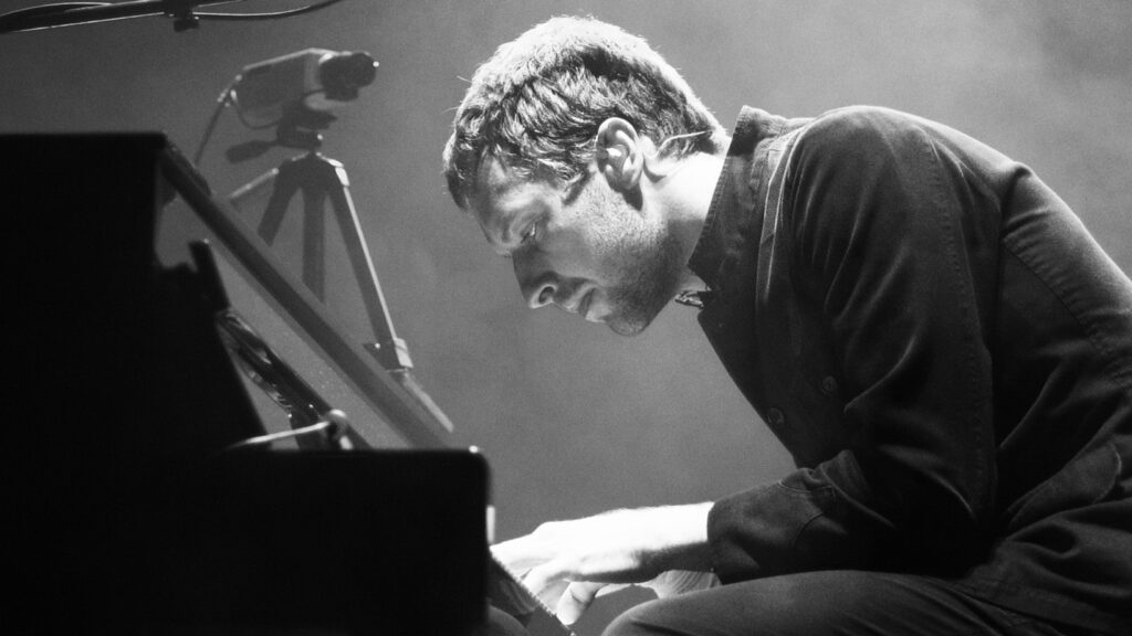 How Many Girlfriends Did Chris Martin Have? Full Relationship History 1 Chris Martin in a black and white photograph, wearing dark clothing and playing piano while leaning over the keys on a stage during a performance.