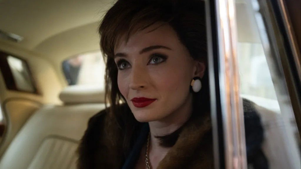 How Many Girlfriends Did Chris Martin Have? Full Relationship History 7 Sophie Turner with short brown hair and red lipstick, wearing white earrings and looking thoughtfully to the side while sitting in a car in a still from Joan.