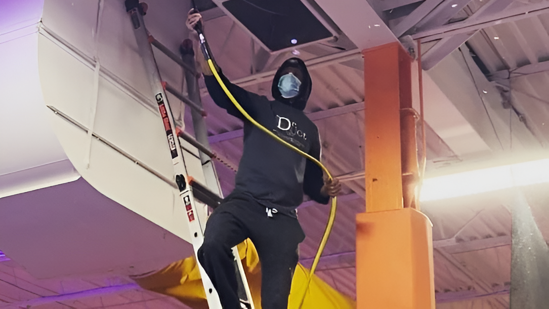 Floyd Roger Myers Jr. in dark clothing and face mask working on ductwork cleaning in a commercial building with exposed ceiling.