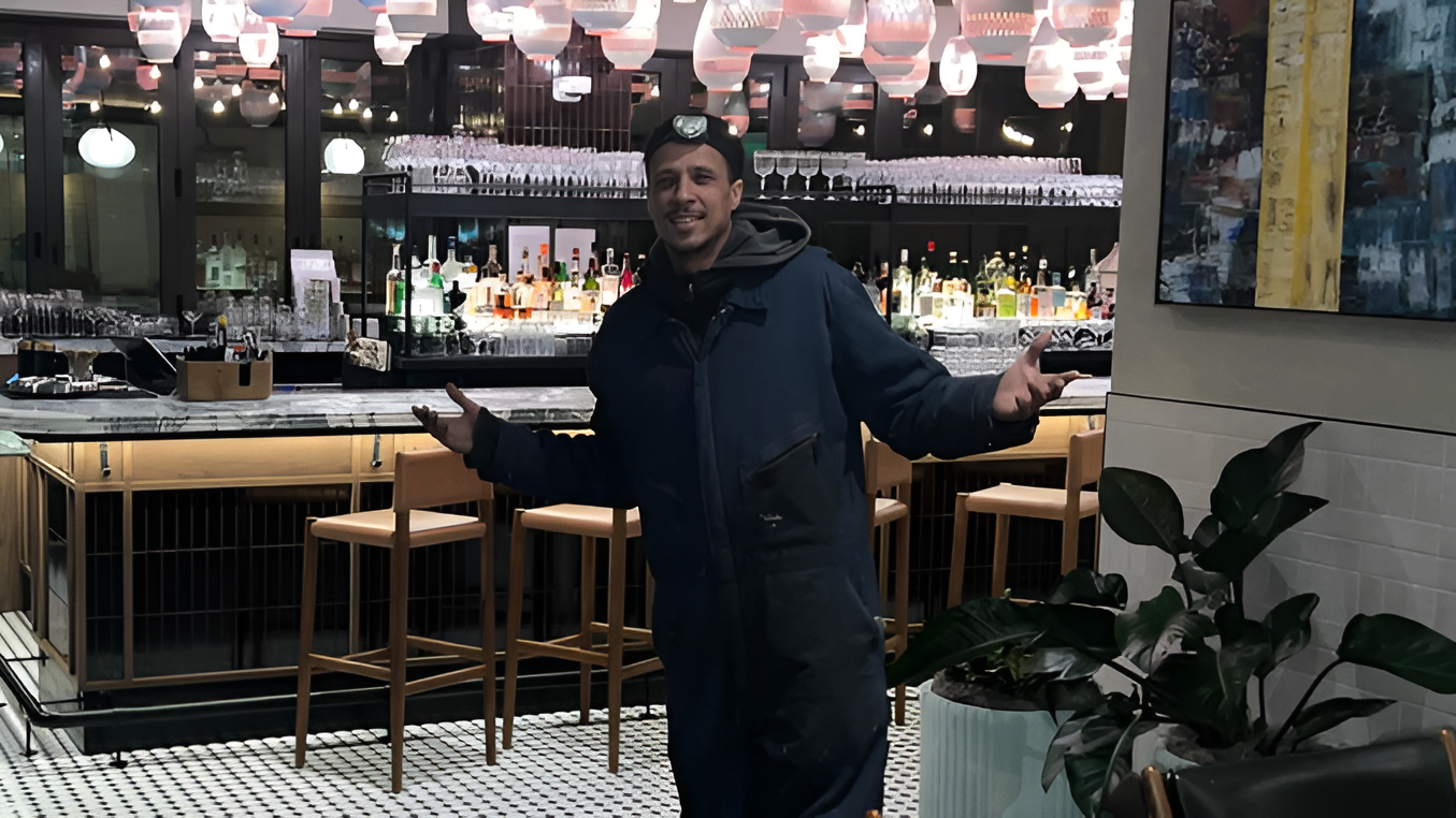 Floyd Roger Myers Jr. smiling in a dark hoodie, standing in a modern restaurant with exposed lighting and contemporary design.