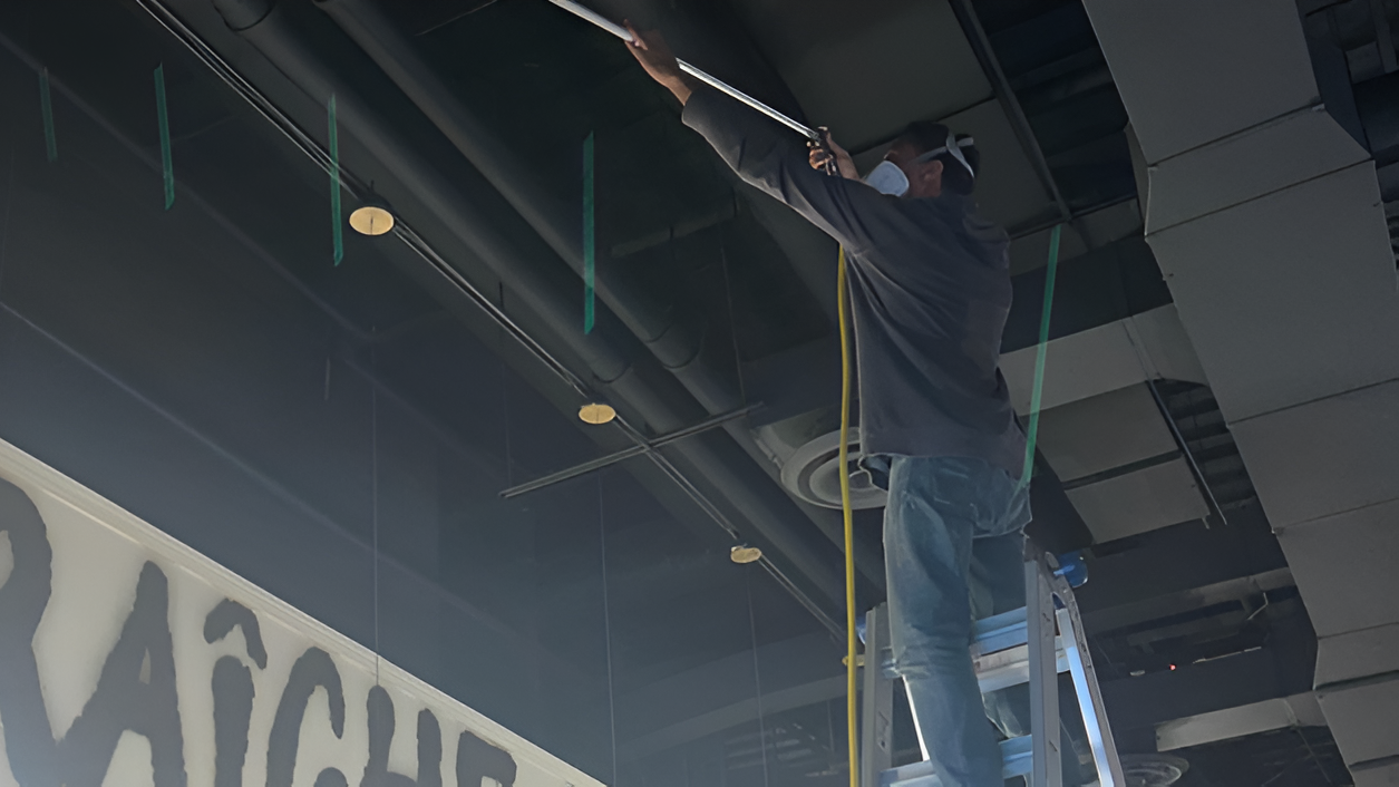 Floyd Roger Myers Jr. working on air duct cleaning, reaching up to clean ceiling ductwork in a commercial space.
