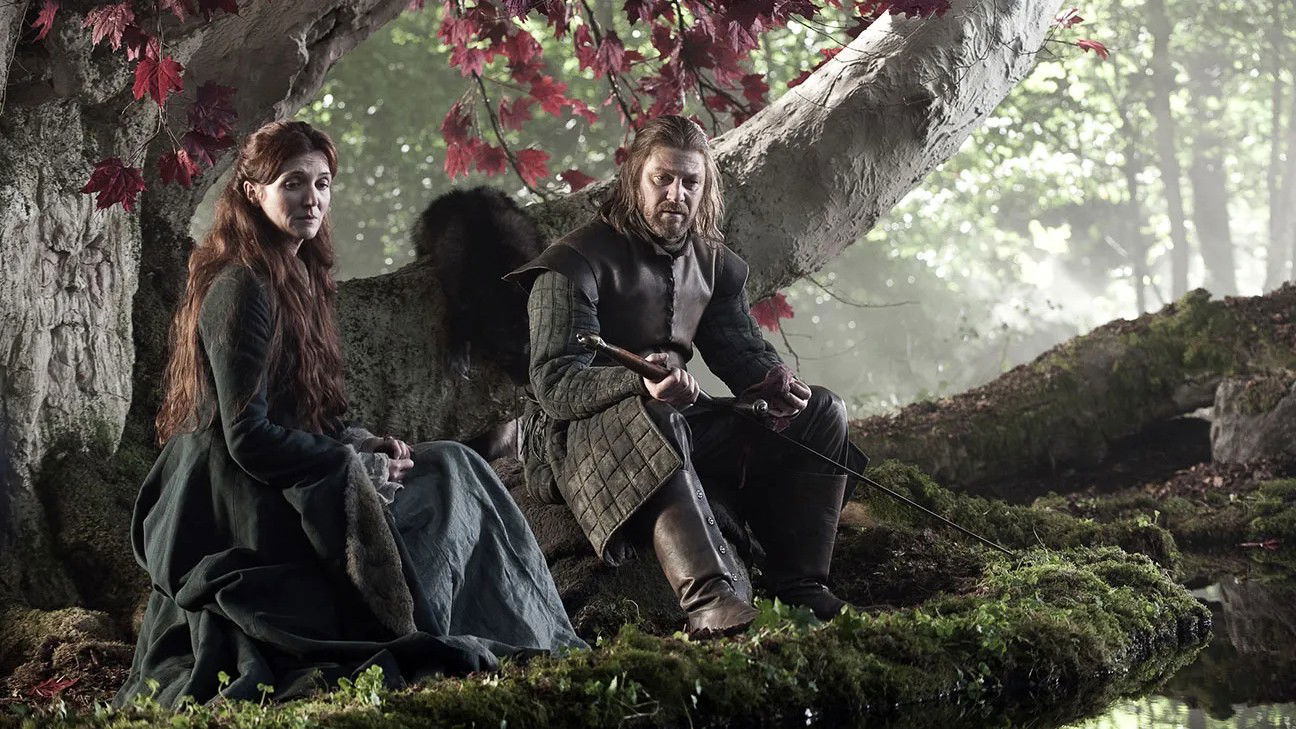 A scene from Game of Thrones showing Catelyn Stark and Ned Stark sitting together under a tree with red autumn leaves, depicting one of their quieter, more intimate moments.