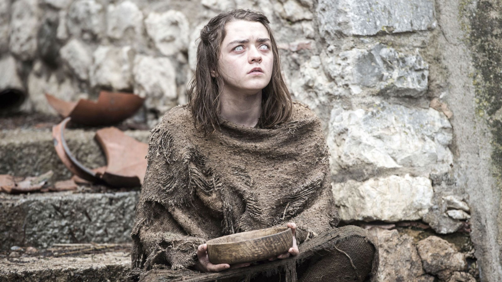 Arya Stark from Game of Thrones, shown as a young girl in rough, tattered clothing sitting against stone ruins, holding a wooden bowl and looking upward with a confused expression.