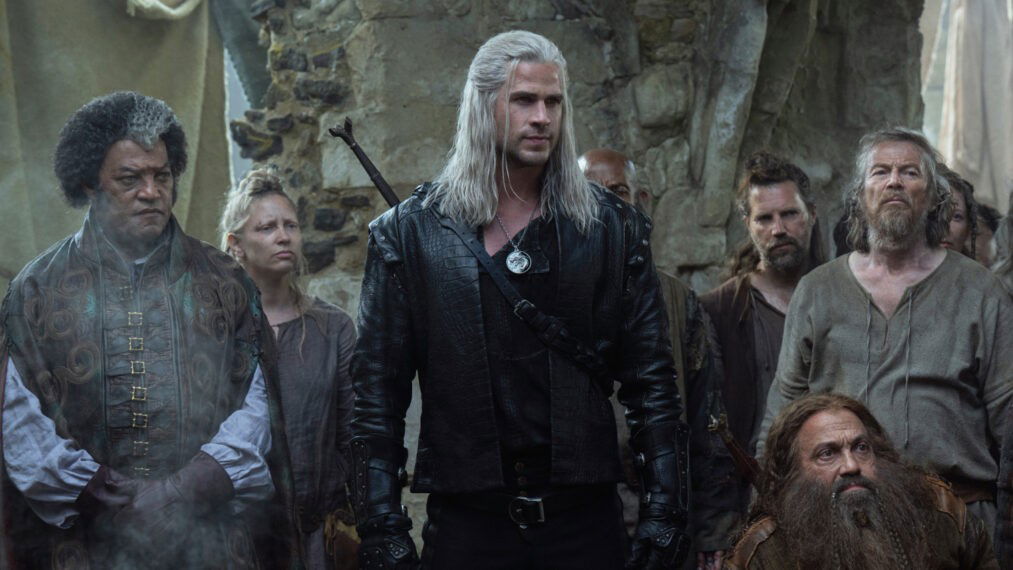 Liam Hemsworth as Geralt in formal witcher attire, standing confidently in a great hall, surrounded by other characters in medieval dress.