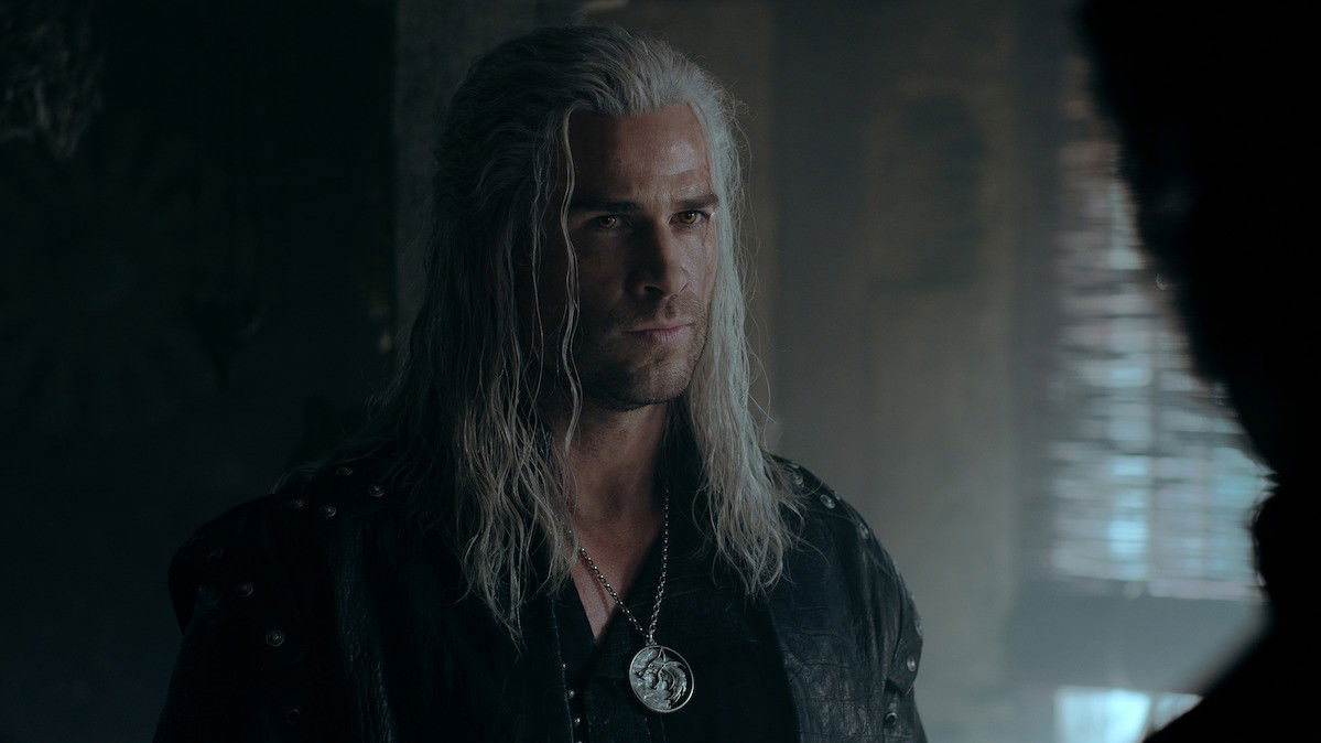 A portrait shot of Liam Hemsworth as Geralt, featuring the white hair and intense gaze, wearing dark leather clothing with the signature wolf medallion visible.