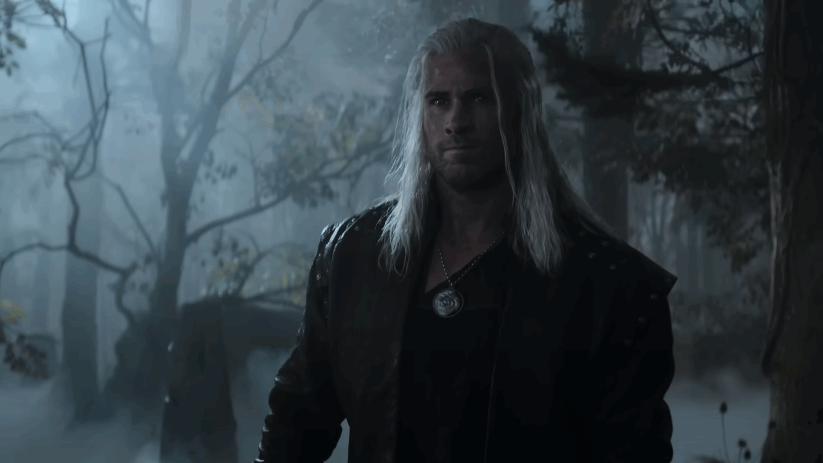 A close-up portrait of Liam Hemsworth as Geralt, showing his serious expression with the characteristic white hair, wearing dark clothing and the wolf medallion against a blurred background.