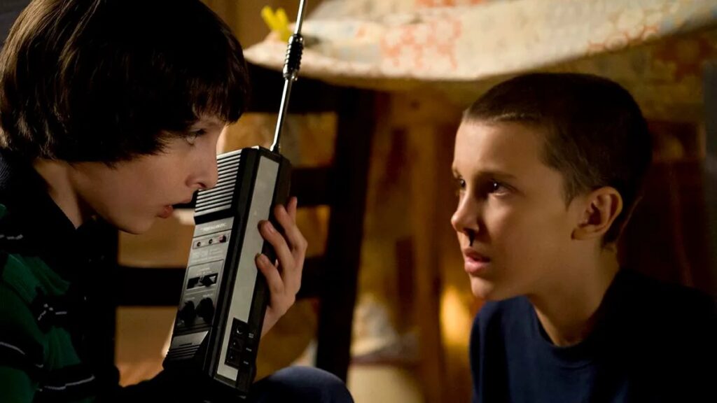 Mike Wheeler (Finn Wolfhard) and Eleven (Millie Bobby Brown) talking while Mike is holding a walkie-talkie radio, both looking concerned in warm indoor lighting.