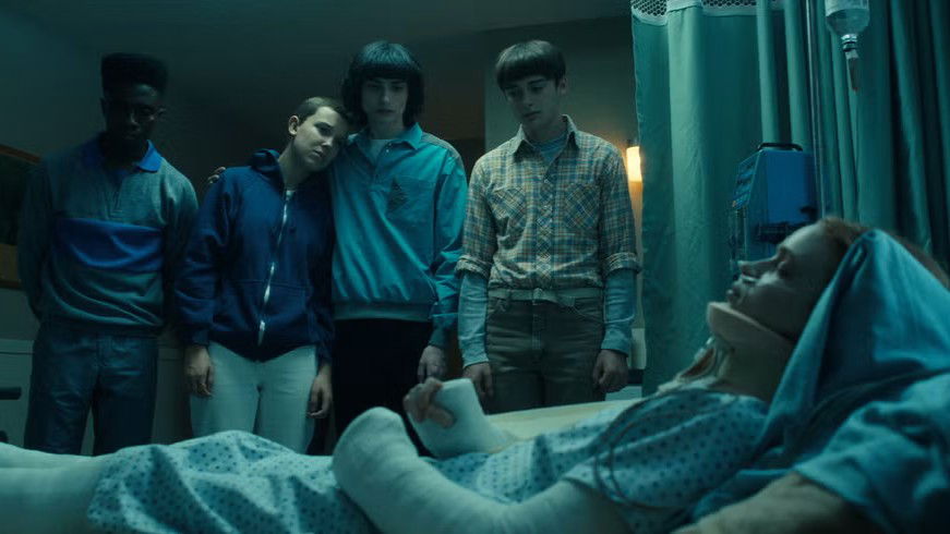 Lucas (Caleb McLaughlin), Mike (Finn Wolfhard), Will (Noah Schnapp), and Eleven (Millie Bobby Brown) standing around a hospital bed in blue-tinted lighting while Max Mayfield (Sadie Sink) is lying in a coma with neck braces and hospital equipment attached to her.